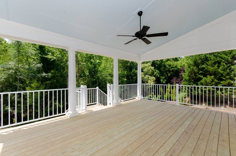 Large wooden deck with white railing, forest backdrop, and a ceiling fan for outdoor living