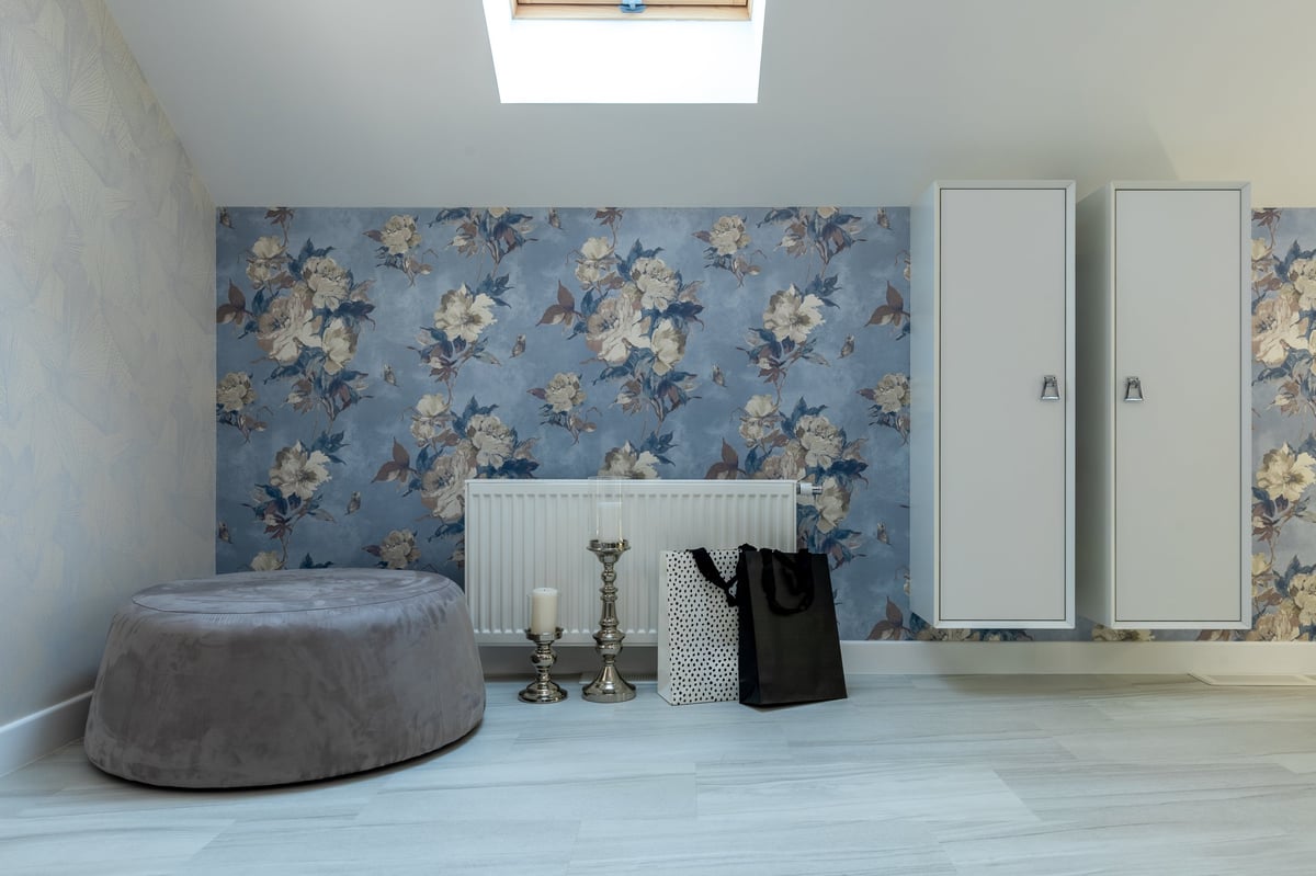 Interior detail of a modern attic bedroom featuring a large gray velvet pouf, elegant silver candle holders, minimalist floating white cabinets, and blue floral pattern wallpaper under a skylight.