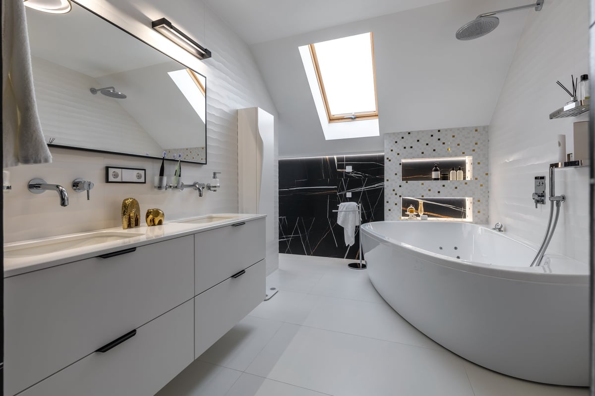 Contemporary bathroom featuring a large white double sink vanity, freestanding soaking tub, gold mosaic accents, and natural light from a skylight.