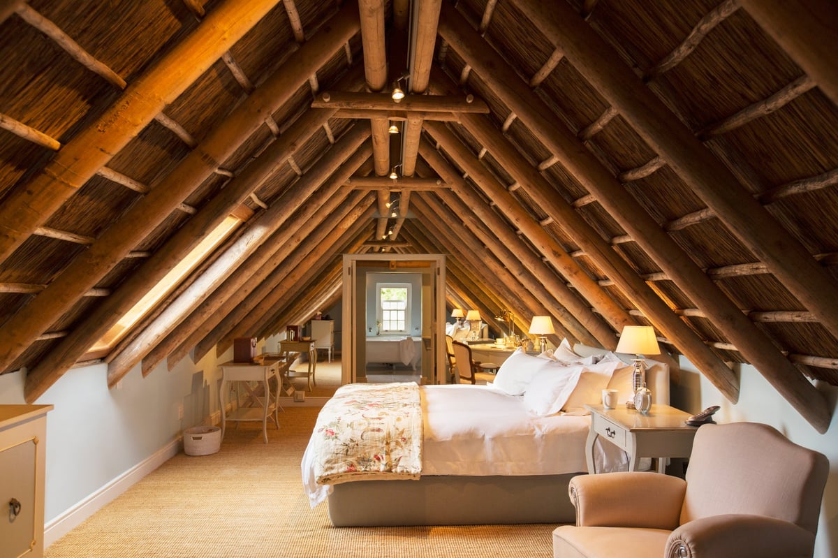 Cozy attic bedroom with exposed wooden beams, a bed with white bedding, armchair seating, warm lighting, and skylight window