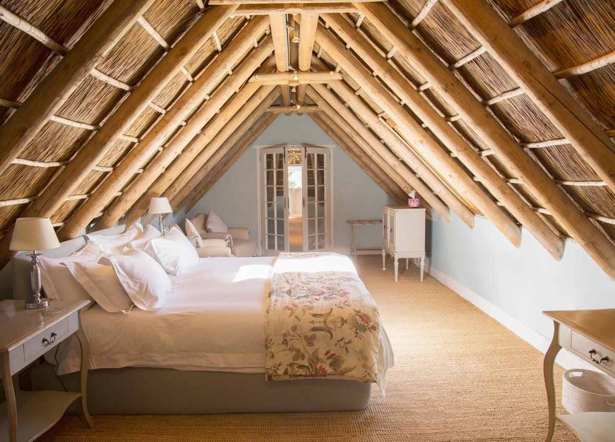 Bright attic bedroom with exposed wooden beams, a white bed with yellow bedding, and a skylight window centered at the far end