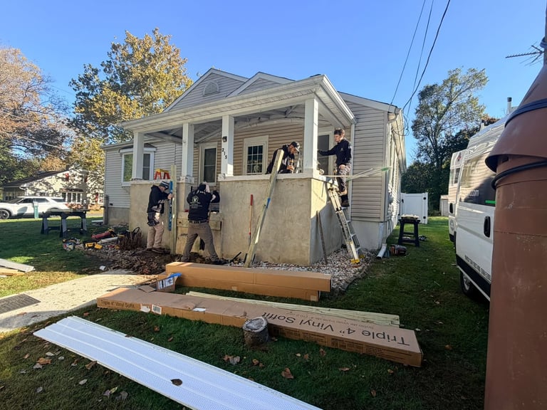 Crew installing exterior siding on a house wall from a ladder with construction materials stacked in the yard