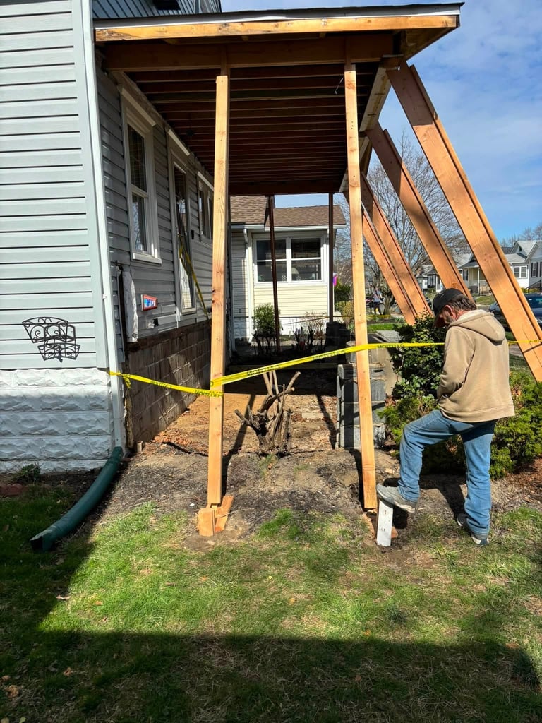 Carpenter in a tan jacket standing behind yellow caution tape under a wooden pergola-like porch framing attached to a house