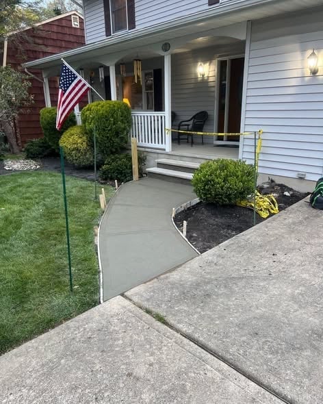 Two-story house front porch with white railing posts, American flag, green shrubs and a concrete walkway to the steps